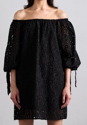 Mujer con un vestido negro de hombros descubiertos con tela de ojales y mangas abullonadas atadas con lazos en los codos, de pie frente a un fondo liso.