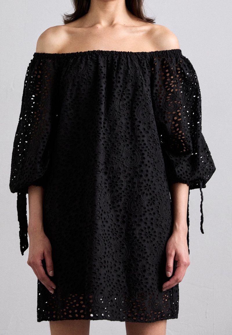 Femme portant une robe noire épaules dénudées en tissu ajouré avec des manches bouffantes nouées par des nœuds aux coudes, debout devant un fond uni.