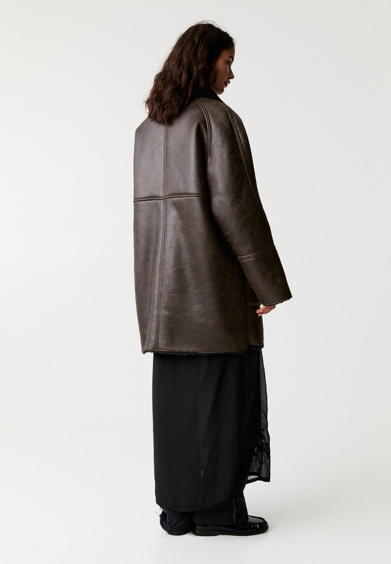 Frau mit langen Haaren, die eine übergroße braune Lederjacke über einem langen schwarzen Rock und schwarzen Schuhen trägt und vor einem weißen Hintergrund steht.