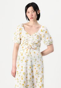 Witte bloemen crop top met gele en groene patronen, met een vierkante halslijn, geplooide details en korte pofmouwen. Gestructureerde stof.