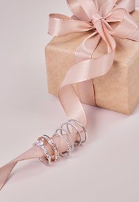 Una pila de anillos de plata y oro con varios diseños, descansando sobre una cinta de satén rosa atada alrededor de una caja de regalo marrón.