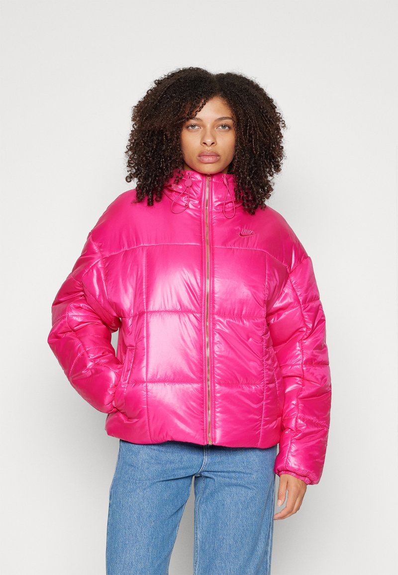 Rosa Puffjacke mit einem hohen Kragen, Reißverschluss und elastischen Akzenten. Verfügt über eine gesteppten Textur und vordere Taschen. Kombiniert mit blauen Jeans.