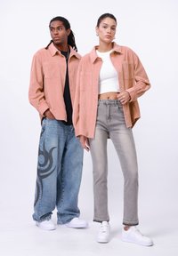Twee modellen in outfits: een lichtroze corduroy shirt, grijze jeans met rafelige randen en witte sneakers. De jeans van het mannelijke model hebben een donker grafisch patroon.