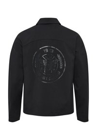 Svart jacka med en glansig, rund logotyp som har ett bi och en lagerkransdesign. Den inkluderar året "1923" och ordet "SPORTSWEAR."