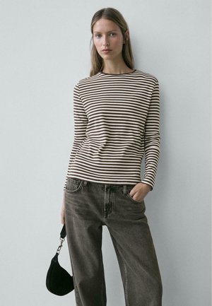 Junge Frau trägt ein braun-weiß gestreiftes Langarmshirt, dunkle Jeans, hält eine kleine schwarze Tasche, mit einer Hand in der Hosentasche.