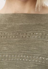 Pull tricot couleur olive avec encolure bateau, présentant des rayures horizontales et des détails décoratifs en ajouré. Le matériau semble doux et texturé.