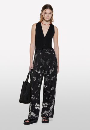 Gilet nero lavorato a maglia con scollatura a V profonda e bottoni, abbinato a pantaloni neri a gamba larga con fantasie floreali bianche. Modella tiene una borsa nera con texture.