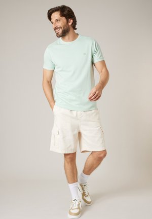 Homme souriant portant un t-shirt vert clair, un short cargo beige, des chaussettes blanches et des baskets beige et blanches, debout devant un fond neutre.