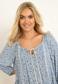 Sininen kukkakuosiset paita, joka on valmistettu kevyestä kankaasta. Siinä on avonainen kaula-aukko, valkoinen solmusepala ja pitkät hihat, jotka antavat rennon istuvuuden.