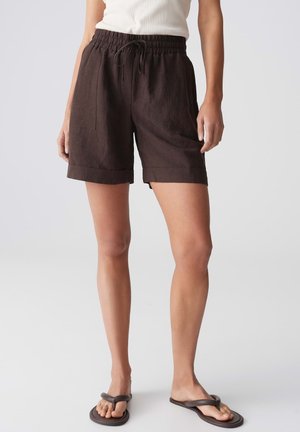 Persoon die donkerbruine shorts met trekkoord en zakken draagt en zwarte teenslippers, staand tegen een effen lichte achtergrond.