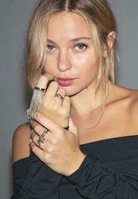 Sølv kæde-frynse ring på den ene hånd, flere ringe på fingrene, neutral hudtone, iført en off-shoulder sort top. Blødt lys.