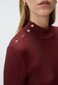 Burgunderfarbener, gerippter Rollkragenpullover mit goldenen Knopfakzenten an der Schulter. Der Stoff wirkt glatt und hat ein vertikales Streifenmuster.
