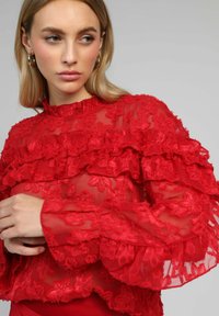 Blouse rouge en voile transparent ornée de broderies florales, de volants superposés, d'un col montant et de manches longues avec des poignets évasés.