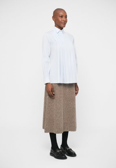 Ženska s obrito glavo, oblečena v svetlo modro plisirano bluzo, rjavo pleteno midi krilo, črne nogavice in črne loafer čevlje z zlatim verigo.