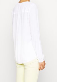 Camicia bianca a maniche lunghe con scollatura arrotondata e orlo dritto sul retro, realizzata in tessuto leggero, indossata con pantaloni gialli chiari.