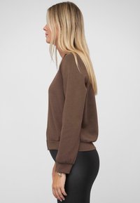 Brauner Sweatshirt mit lockerer Passform, rundem Halsausschnitt und langen Ärmeln. Gerippter Saum an der Taille. Getragen mit schwarzen Leggings.