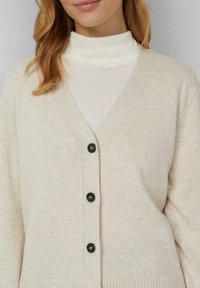 Beżowy kardigan z dzianiny, z dekoltem w kształcie litery V, pięcioma czarnymi guzikami i prążkowanymi mankietami. Pod spodem biały golf.