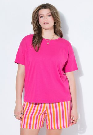 Frau trägt ein hellrosa kurzärmeliges Shirt und pink-orange-weiße vertikal gestreifte Shorts, steht vor einem weißen Hintergrund.