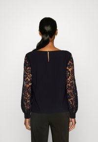Marinebluse mit transparenten langen Spitzenärmeln. Verfügt über einen geraden Saum und eine kleine Öffnung mit Schlüsselloch hinten. Glattes Material mit strukturierten Spitzelementen.