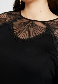 Schwarzes Oberteil mit transparenter schwarzer Spitze im wellenförmigen Muster am Ausschnitt, glatter Textur und rundem Kragen.