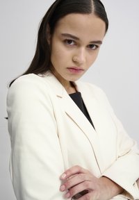 Bežový tailored blazer s hladkým povrchem a strukturovanými ramenními vycpávkami. Obsahuje klopové límce a jednořadé zapínání na knoflík.