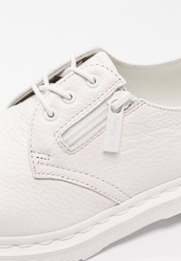 Chaussure en cuir blanc avec surface texturée, lacets blancs et fermeture éclair latérale étiquetée « Dr. Martens » sur fond blanc.