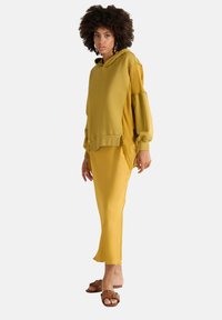 Haut à capuche oversize couleur moutarde avec un tissu texturé, des manches ballon et des détails en franges, associé à une jupe midi jaune assortie et des sandales à glisser brunes.