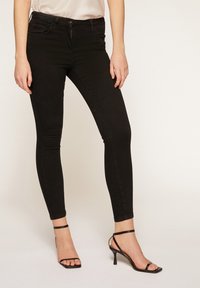 Jeans negros pitillo con cintura de talle medio, con cierre de botón y cremallera. Combinados con tacones negros de tiras, textura suave y forma ajustada.