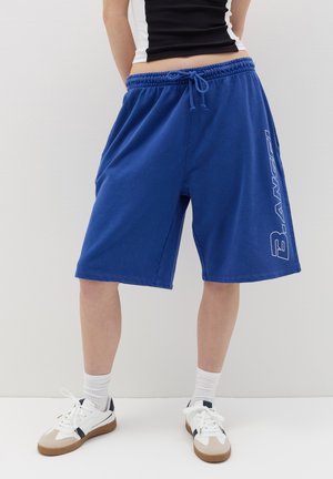 Blue cotton shorts with an elastic waistband and drawstring. Features a white logo printed on the side. Worn with white sneakers and socks.