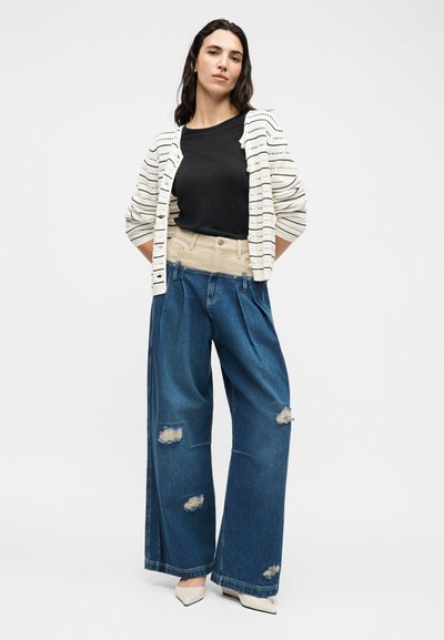 Femme debout portant un haut noir, un cardigan blanc rayé, un jean bleu taille haute à jambes larges avec une ceinture beige, et des chaussures blanches pointues.