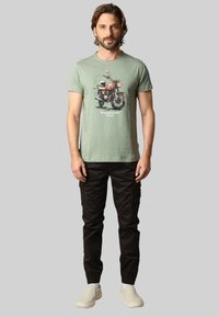 T-shirt vert clair à manches courtes avec un graphique de moto, associé à un pantalon cargo noir et des baskets claires.