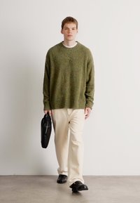 Maglione texture verde oliva con colletto rotondo, abbinato a pantaloni color crema e scarpe slip-on nere. La modella porta una borsa nera.