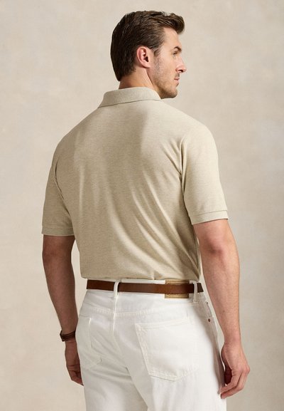 Mies, joka käyttää beigeä lyhythihaista poolopaitaa ja valkoisia housuja ruskean vyön kanssa, seisoo selin yksiväristä beigeä taustaa vasten.