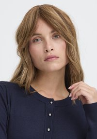 Marineblauwe cardigan met zilveren knopen, geribbelde kraag en manchetten. Het model heeft golvend bruin haar en is van schouders omhoog gefotografeerd tegen een lichte achtergrond.