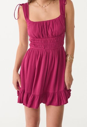 Robe sans manches magenta avec corsage froncé, bretelles nouées aux épaules, ourlet à volants, portée avec un collier chaîne doré et un large bracelet doré.