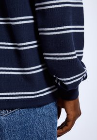 Navy striped sweater featuring white horizontal stripes, textured fabric, ribbed cuffs, worn with blue denim jeans, showing a casual fit.