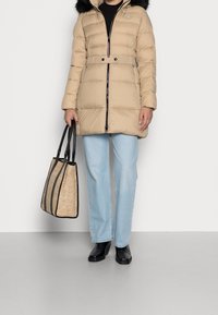Beżowa kurtka puchowa z futrzanym kołnierzem, czarnym zamkiem błyskawicznym i paskiem. Połączona z jasnoniebieskimi dżinsami o szerokich nogawkach oraz czarno-beżową torbą typu tote.