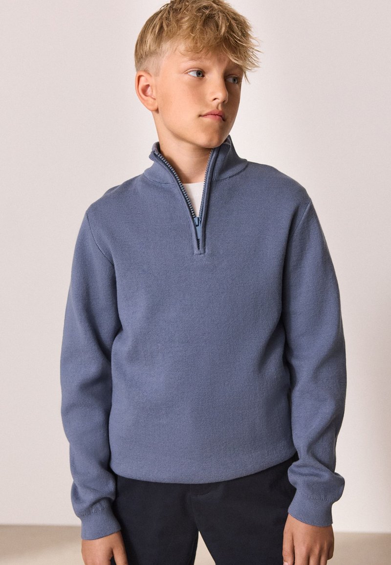 Svetlomodrý sveter so zipsom pri krku z mäkkého pleteného materiálu, s dlhými rukávmi a voľným strihom, v kombinácii s tmavými nohavicami.