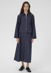 Jeansowa kurtka z zamek błyskawicznym z przodu, długimi rękawami i kołnierzykiem, w połączeniu z pasującą długą spódnicą, wszystko w głębokim niebieskim odcieniu. Czarne buty dopełniają całość.