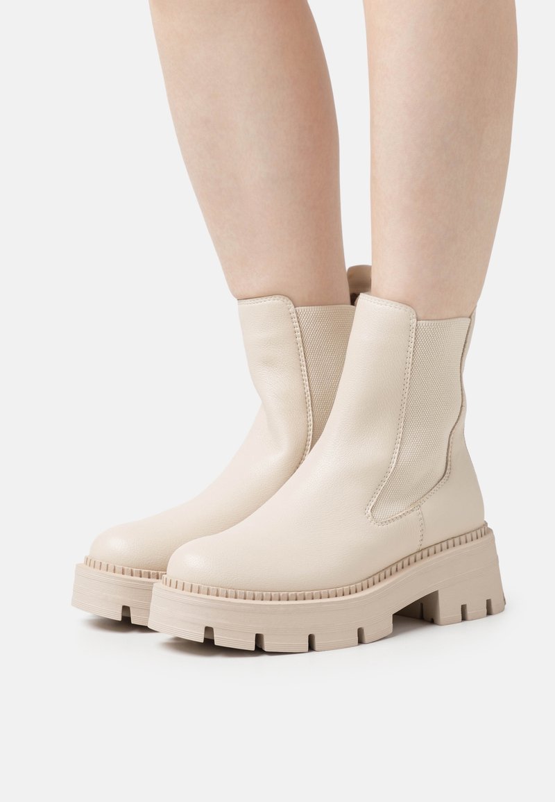 Jambes portant des bottines à plateforme épaisses beiges avec des panneaux latéraux élastiques et des semelles en caoutchouc texturées sur un fond clair.