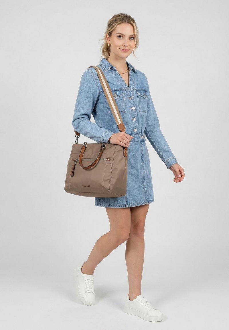 Joven mujer con vestido vaquero y zapatillas blancas, llevando un bolso beige de hombro con asas marrones y una correa a rayas, posando sobre un fondo blanco.