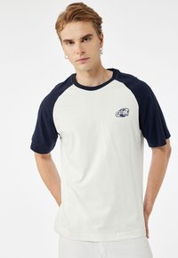 Baumwoll-T-Shirt mit einem cremefarbenen Körper und navy-blauen Raglanärmeln. Es hat einen runden Halsausschnitt und ein Logo auf der linken Brust. Glatte Textur.