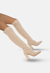 Botas de punto beige hasta la rodilla con un acabado texturizado, punta cuadrada y tacón grueso. El diseño incluye un caño ajustado y acentos lisos.