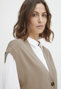 Gilet a maglia beige con scollatura a V, maniche ampie e chiusura con un solo bottone, indossato sopra una camicia bianca con bottoni. Tessuto liscio.