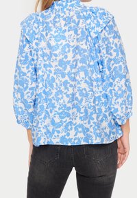 Blus med blå blommönster och volangdetaljer vid axlarna, tillverkad av lättviktig tyg, med långa, sammandragna ärmar. Matchad med mörka jeans.