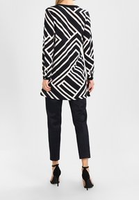Schwarz-weiße Tunika mit geometrischem Muster, lange Ärmel. Kombiniert mit strukturierten, marineblauen Hosen und schwarzen High Heels. Einfache runde Ausschnitt.