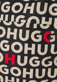 Tissu noir avec motif répétitif de texte beige "HUGO" et lettres rouges occasionnelles, avec détails des coutures et des poches.