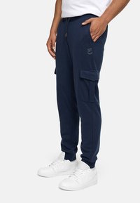 Marineblaue Cargo-Jogginghosen aus weichem Material mit Seitentaschen, elastischem Bund und Logodetail am Oberschenkel. Kombiniert mit weißen Sneakern.