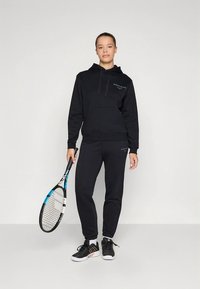 Sweatshirt preta com capuz e calças de fato de treino com detalhes de marca, apresentando um corte descontraído e tecido texturizado. Equipado com uma raquete de ténis.