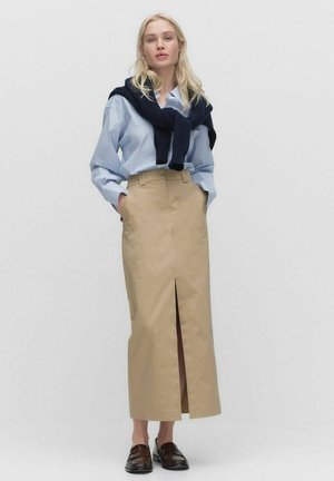 Jupe beige taille haute avec fente à l'avant, assortie à une chemise boutonnée rayée bleu clair et un pull navy noué autour des épaules.